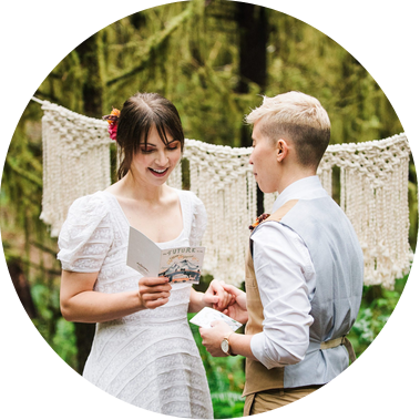 An eloping couple reads their vows in front of a macrame backdrop that's been strung between two trees in a forest