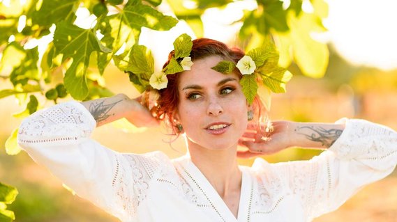 A young, tattooed bride lifts her arms above her head as she stands in the shade of a fig tree