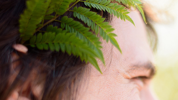 A close view of a fellow's face; we see a single green fern tucked behind his right ear