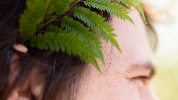 A close view of a fellow's face; we see a single green fern tucked behind his right ear