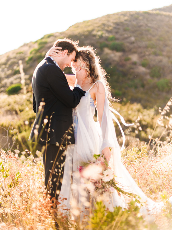 A bride and groom embrace in the warm sun of late summer, surrounded by golden hills