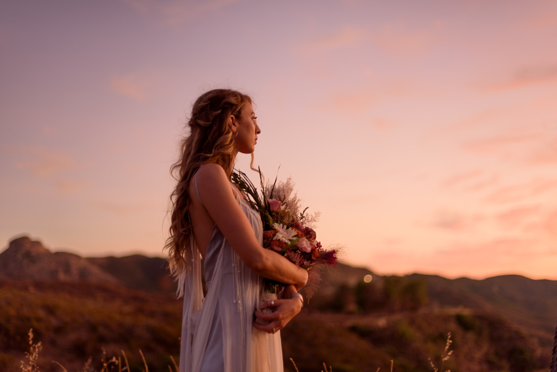 A bride holds her bouquet and gazes out at the fading sunset, colored pink by the last rays of sunlight
