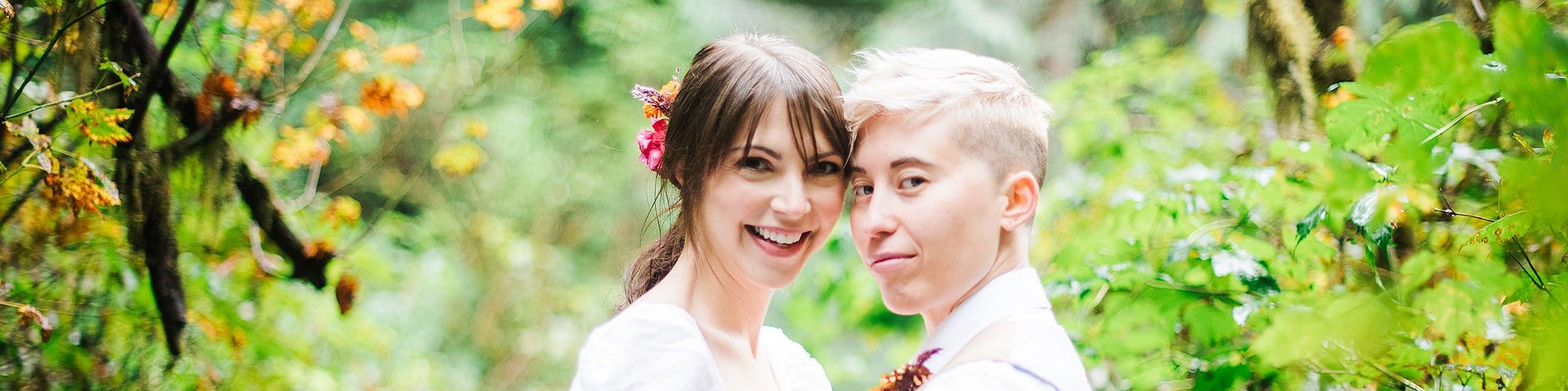 An eloping couple looks at us, smiling, their heads together; standing on a narrow hiking path, they are surrounded by wild plants