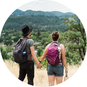 A man and a woman stand on the side of a hiking trail, and look out and the tree-filled view below them
