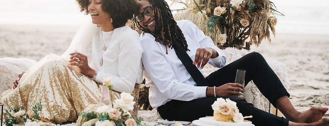 While picnicking and enjoying champagne, the bride and groom lean back-to-back, look out at the landscape, and laugh