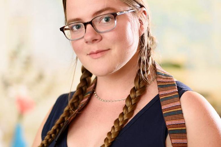 A portrait of the photographer, Kate Waterman Rose. Standing indoors, she wears glasses and her hair is long and braided; she looks out at the viewer thoughtfully and wears a camera strap around her neck