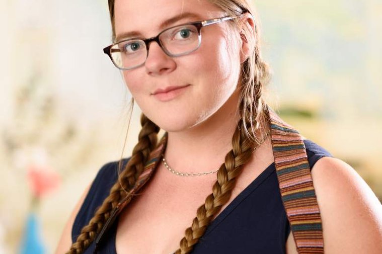 A portrait of the photographer, Kate Waterman Rose. Standing indoors, she wears glasses and her hair is long and braided; she looks out at the viewer thoughtfully and wears a camera strap around her neck