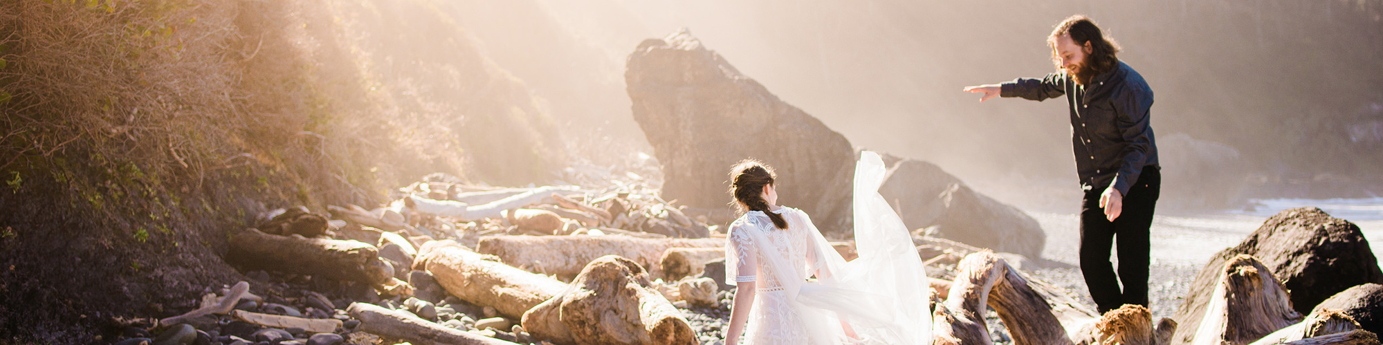 A bride and groom frolic on a Northern California beach; the bride jumps down from a pile of driftwood, her veil flowing behind her