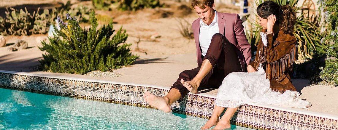 a newlywed couple sits on the edge of their AirBnB pool as they dip their feet into its turquoise waters