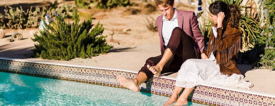 a newlywed couple sits on the edge of their AirBnB pool as they dip their feet into its turquoise waters