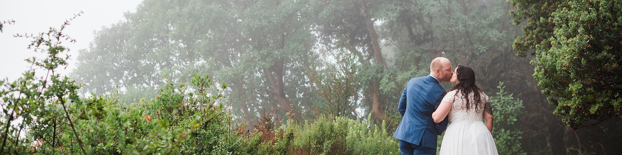 A bride and groom kiss as they walk down a misty forest path