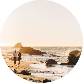 An engaged couple walks calmly along a rocky beach as the sun begins to set