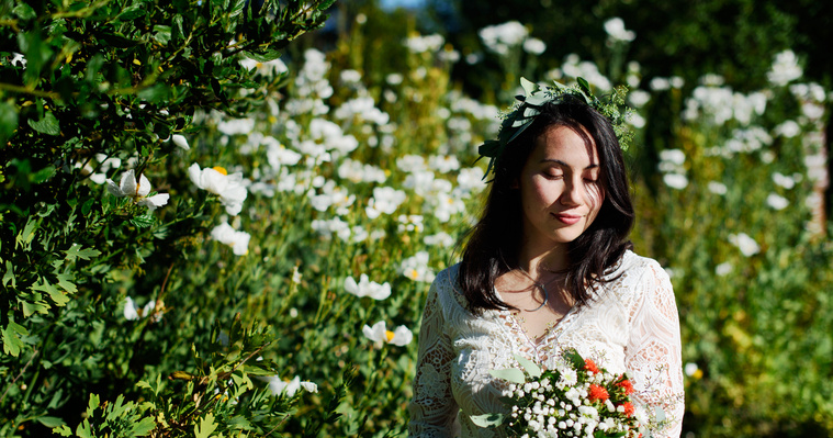 A bride smiles subtly, her eyes downcast, as she holds her bouquet in front of a thicket of wildflowers