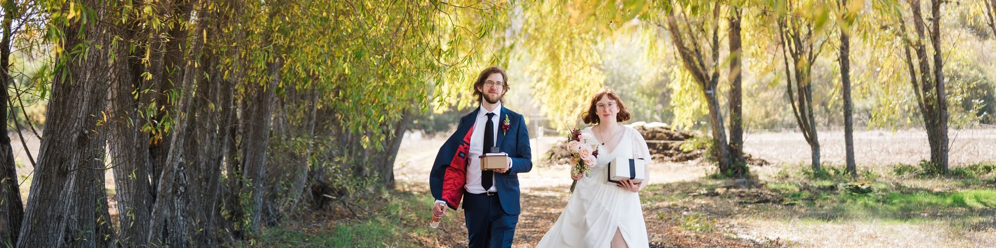 An eloping bride and groom walk down a windy lane under autumn trees, carrying a cake box