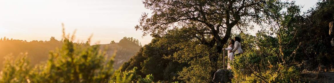 Two brides stand together on a rocky ledge overlooking a mountain sunset