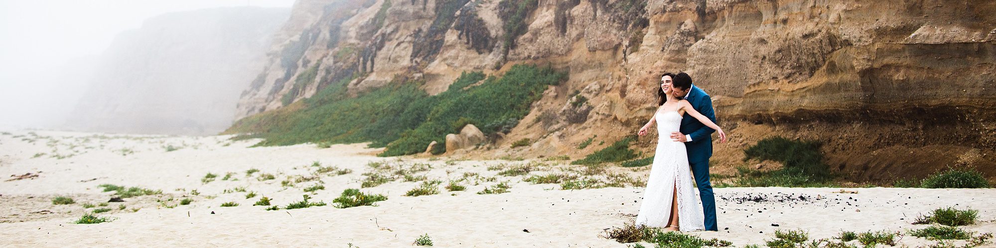 A groom kisses his bride's neck as they stand together on a foggy beach below striated red bluffs