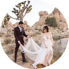 A groom holds his bride's train as they walk through the desert at Joshua Tree National Park
