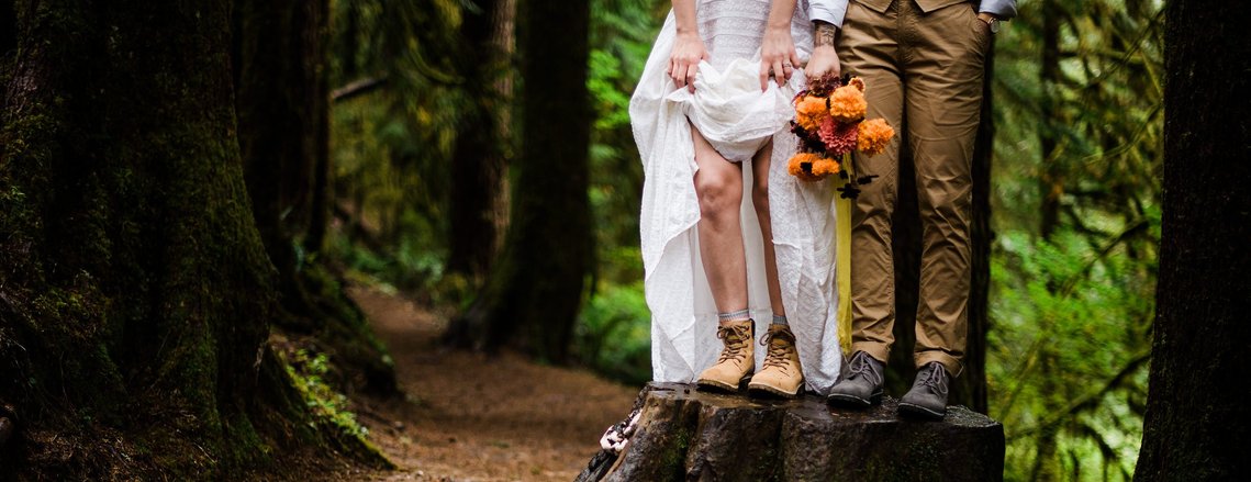 An eloping couple stand on a large tree stump in the forest; the bride lifts the skirt of her gown to show her rugged hiking shoes