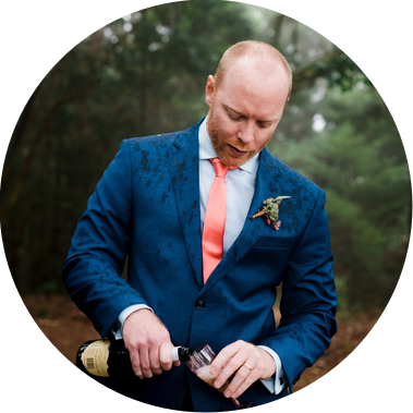 A groom pours champagne into his glass; drops of water speckle his shoulders as he and his bride elope under a canopy of trees on a rainy evening
