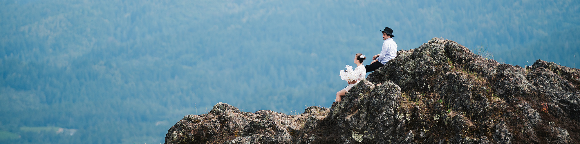 An eloping bride and groom perch on a large rock formation, looking out at the hazy blue mountains in the distance