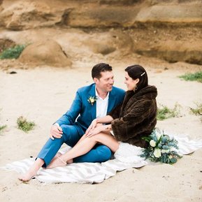 A bride and groom cuddle together on a sandy beach, with golden bluffs rising up behind them
