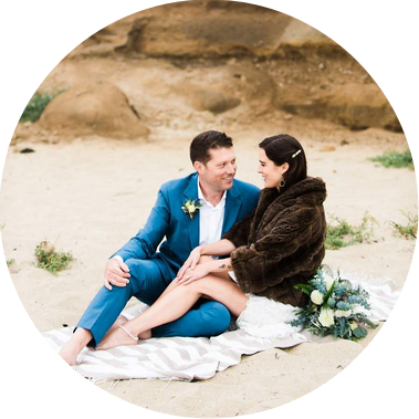 A bride and groom sit on the beach at Half Moon Bay, smiling and cuddling on a beach blanket