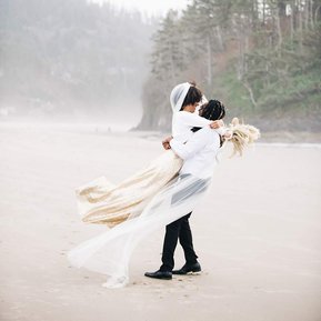 At their elopement on an Oregon Coast  beach, a groom lifts up his bride and twirls her around, her veil trailing in the wind