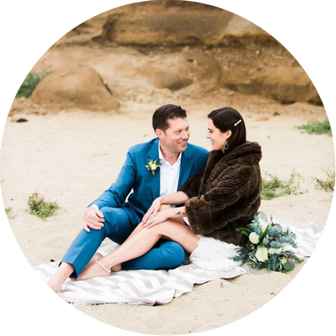 San Francisco Elopement Photography Packages: A happy couple in wedding clothes sits on a beach blanket in the sand, snuggling together and laughing