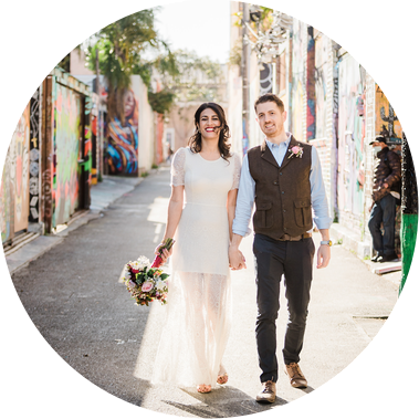 A bride and groom walk together down a street in the San Francisco Mission District, surrounded by colorful murals