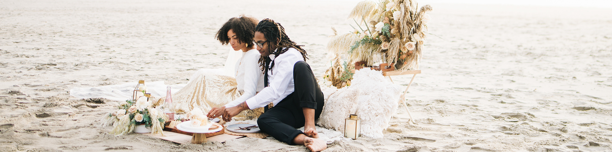 An eloping bride and groom sit on a beach blanket surrounded by elaborate florals, feasting on charcuterie, champagne, and cake