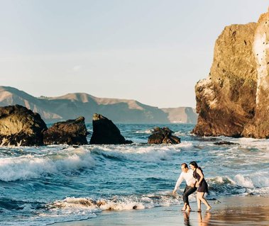 a couple celebrating their engagement run and play in the surf at a rocky California beach