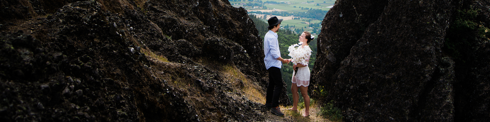 A bride and groom face each other in a valley between two large, dark rock formations