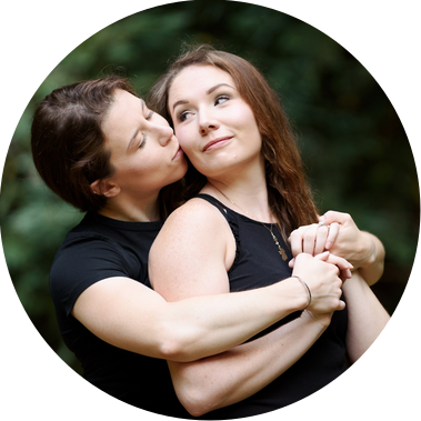 Celebrating their engagement, two women embrace in a redwood forest; one kisses the other from behind