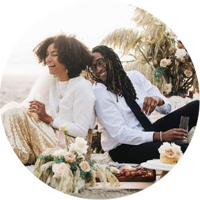 An eloping bride and groom lean back to back, sitting on a beach blanket and surrounded by elaborate florals