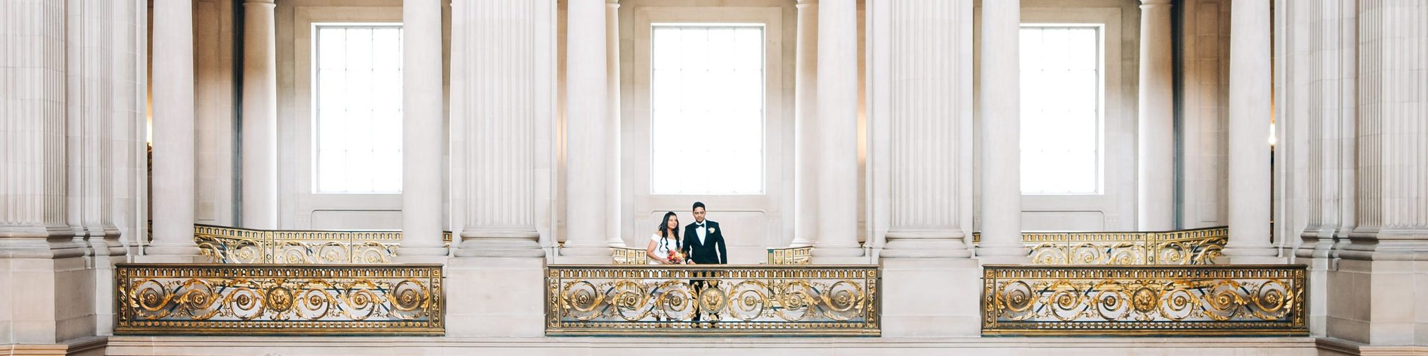Dwarfed by the grand architecture of San Francisco City Hall, a bride and groom look out across the space from a columned balcony