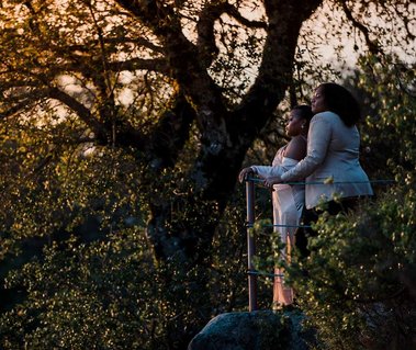 Two brides stand below an oak tree, looking out at the glowing sunset