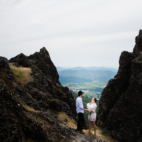 A spunkily-dressed bride and groom face each other on a hilltop between a pair of large, dark rock formations