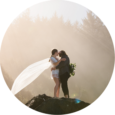 A bride and groom stand atop a large boulder, holding each other and kissing. The bride's cape-veil streams out behind her, held aloft by the breeze
