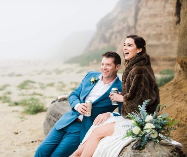A bride and groom sit together on a driftwood log as their enjoy their favorite beers after an elopement ceremony