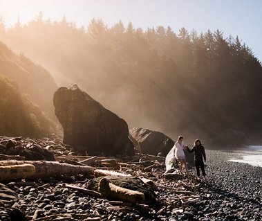 An eloping couple walks hand-in-hand down a rocky Northern California beach, the mountains rising in the morning fog above them