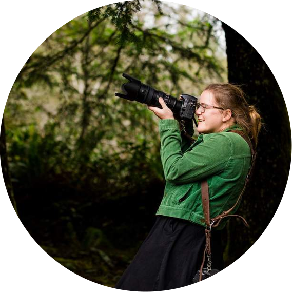 Kate Waterman Rose, elopement photographer extraordinaire, smiles and leans back as she raises a large camera to her eye. She is surrounded by forest