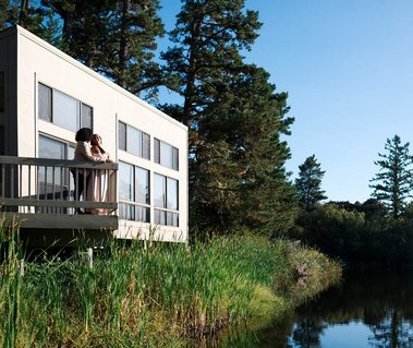 A loving queer couple stands on the deck of a small building overlooking a pond; they cuddle, looking out at the natural world around them