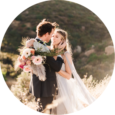 A groom holds his bride as they look into each others' eyes on a golden, grassy hillside