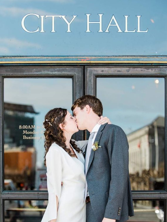 A bride and groom kiss passionately below a sign reading 