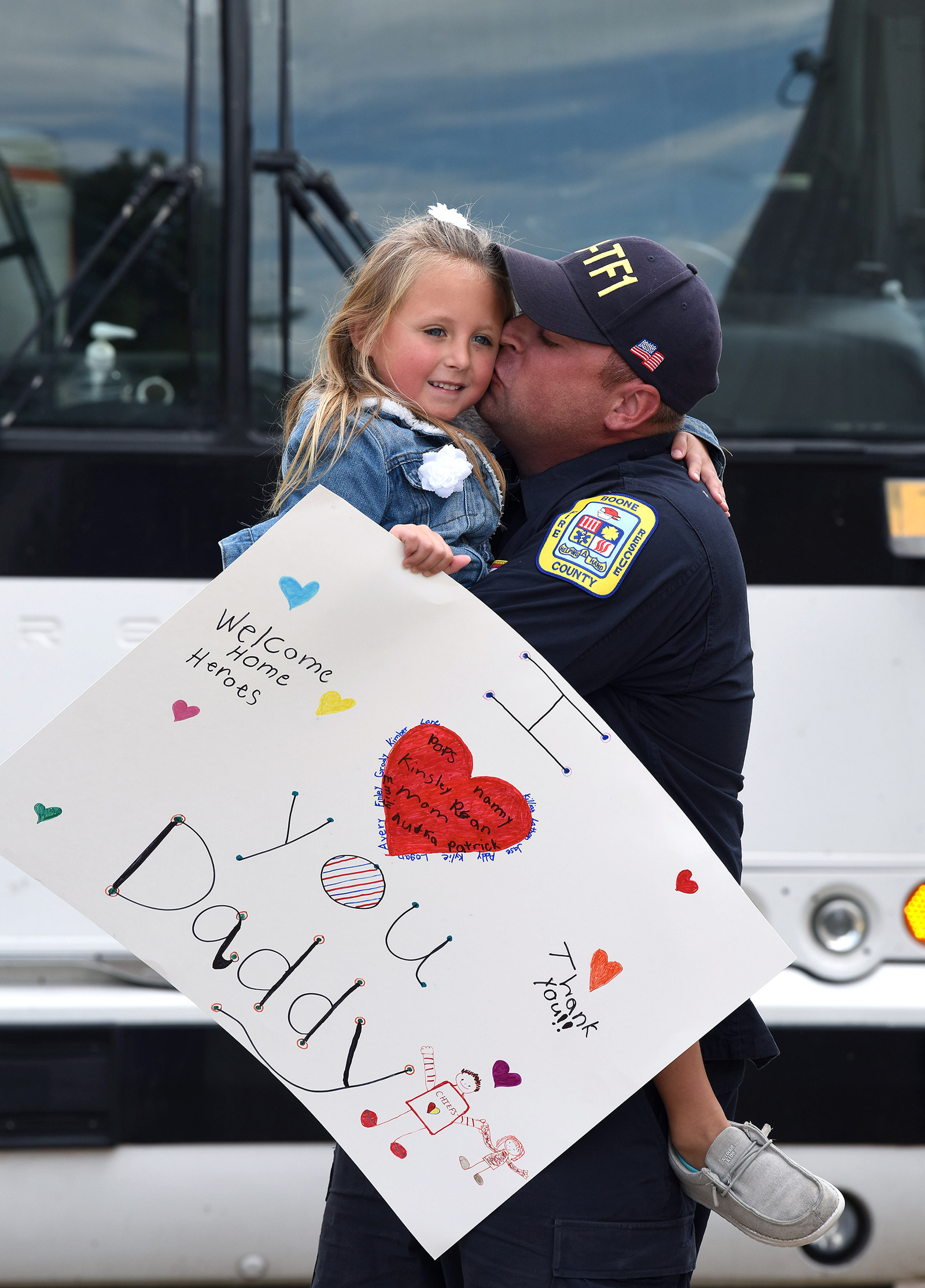A little girl holding a sign that reads 'I love you Daddy' is held by her father as he gives her a kiss on the cheek.