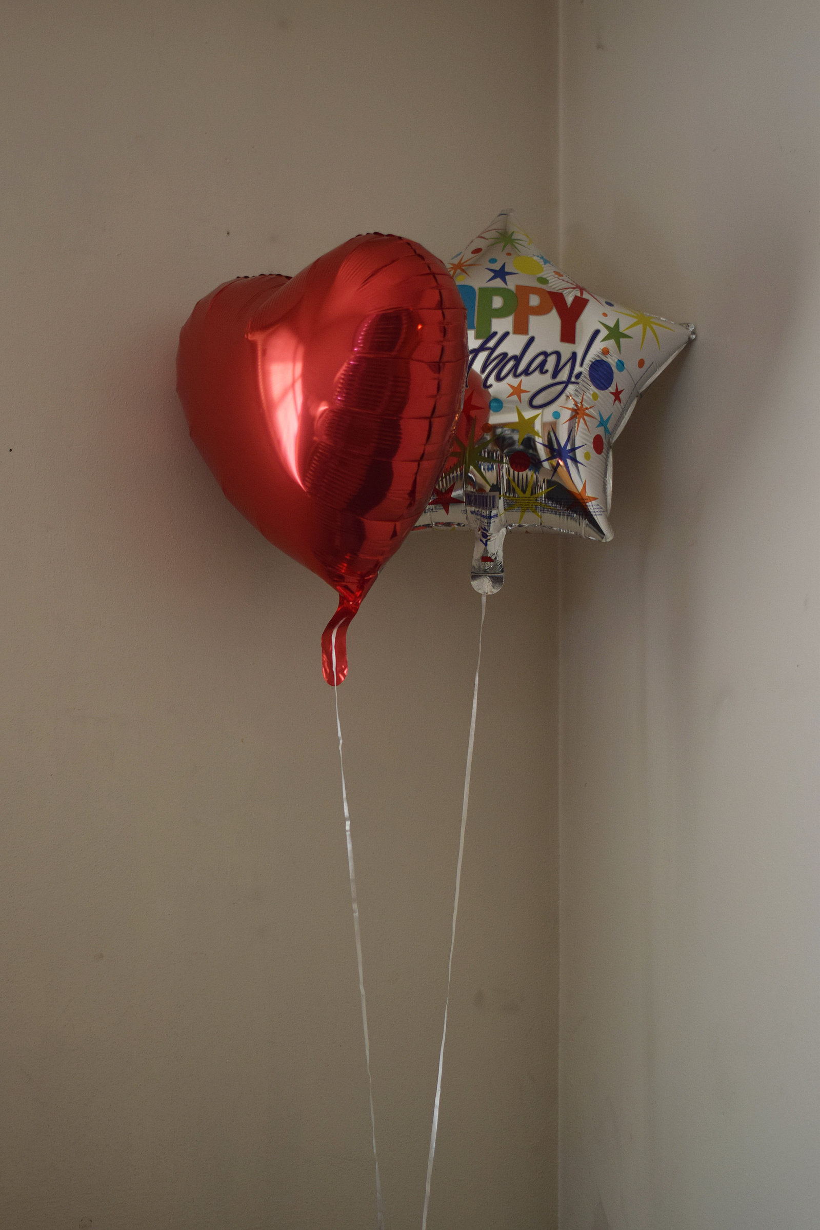 A red heart-shaped balloon and a star-shaped balloon that reads 'Happy Birthday!' rest in the corner of a room.