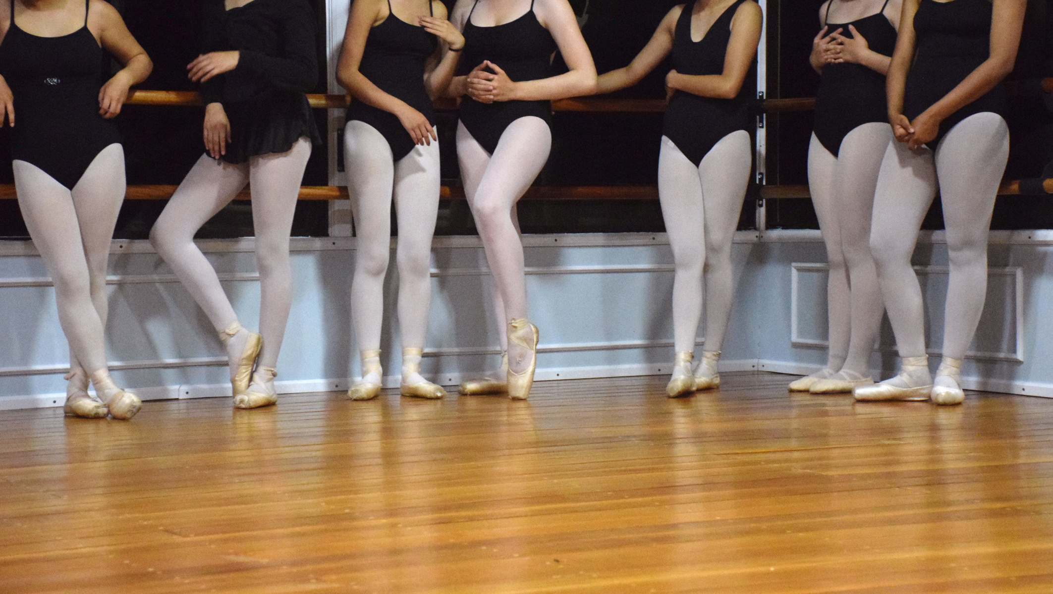 Seven young ballet dancers stand at the edge of their studio and relax before they take turns dancing across the floor.