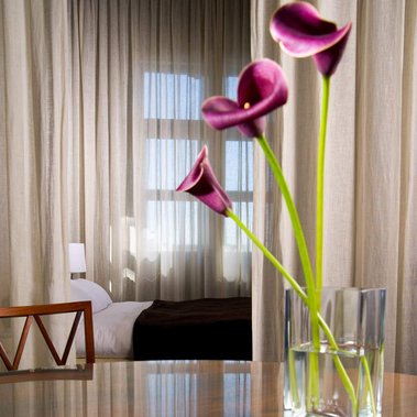 Chambre Hôtel St-Paul, Vieux-Montréal avec vase de fleurs au premier plan et lit fenêtre et rideaux derrière