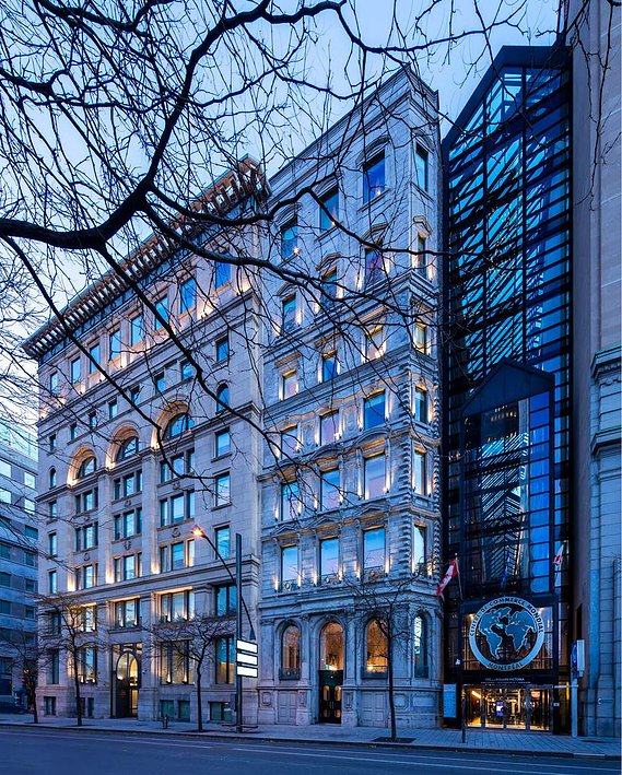 Centre de Commerce mondial à Montréal, photograpjié au lever du jour, avec les branches d'arbres dénudées au premier plan, couleur froides qui contrastent avec la chaleur de l'éclairage intérieur,  on aperçoit toute la façade et ses sept étages