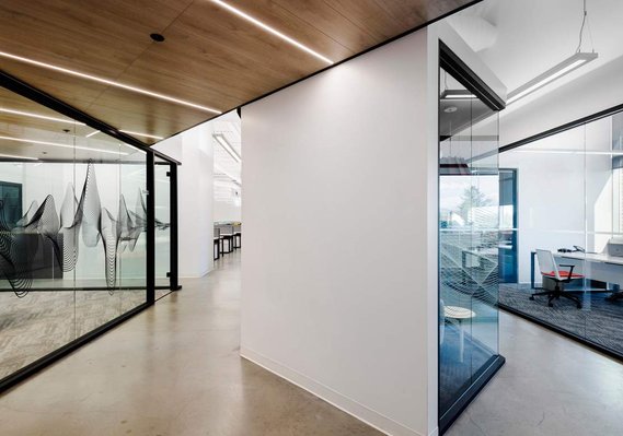 Photo architecturale de l'aménagement d'un bureau montrant la salle de conférence vitrée, une cloison blanche au centre et le corridor menant à des bureaux du côté opposé